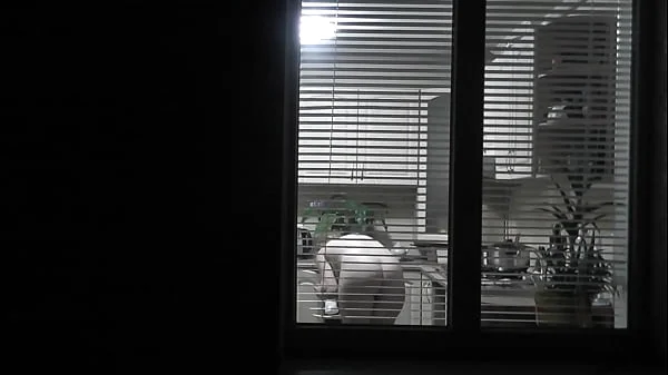Peeping. Voyeur. Neighbor pervert voyeur in evening on street looks out window as naked sexy neighbor in kitchen prepares dinner for her husband. Naked in public. Naked at home. Family.  Outdoor